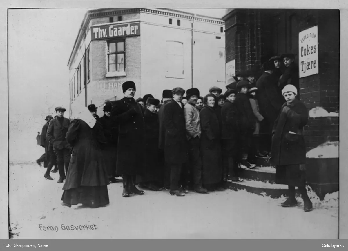 Mennesker i kø foran Gassverket, skilt med "Expedition Cokes Tjære", bygård i bakgrunn med skilt "Thv. Gaarder". Menneskene er godt kledd, vinter, snø på bakken. Ankertorget, Hausmanns gate.