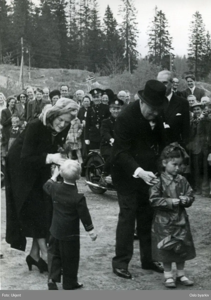 Statsminister Winston Churchill og Mrs. Churchill mottar blomster av barn ved besøk i Oslo 13. mai 1948. Feststemte tilskuere med norske flagg i bakgrunnen. Bildet er trolig tatt på Frognerseteren.