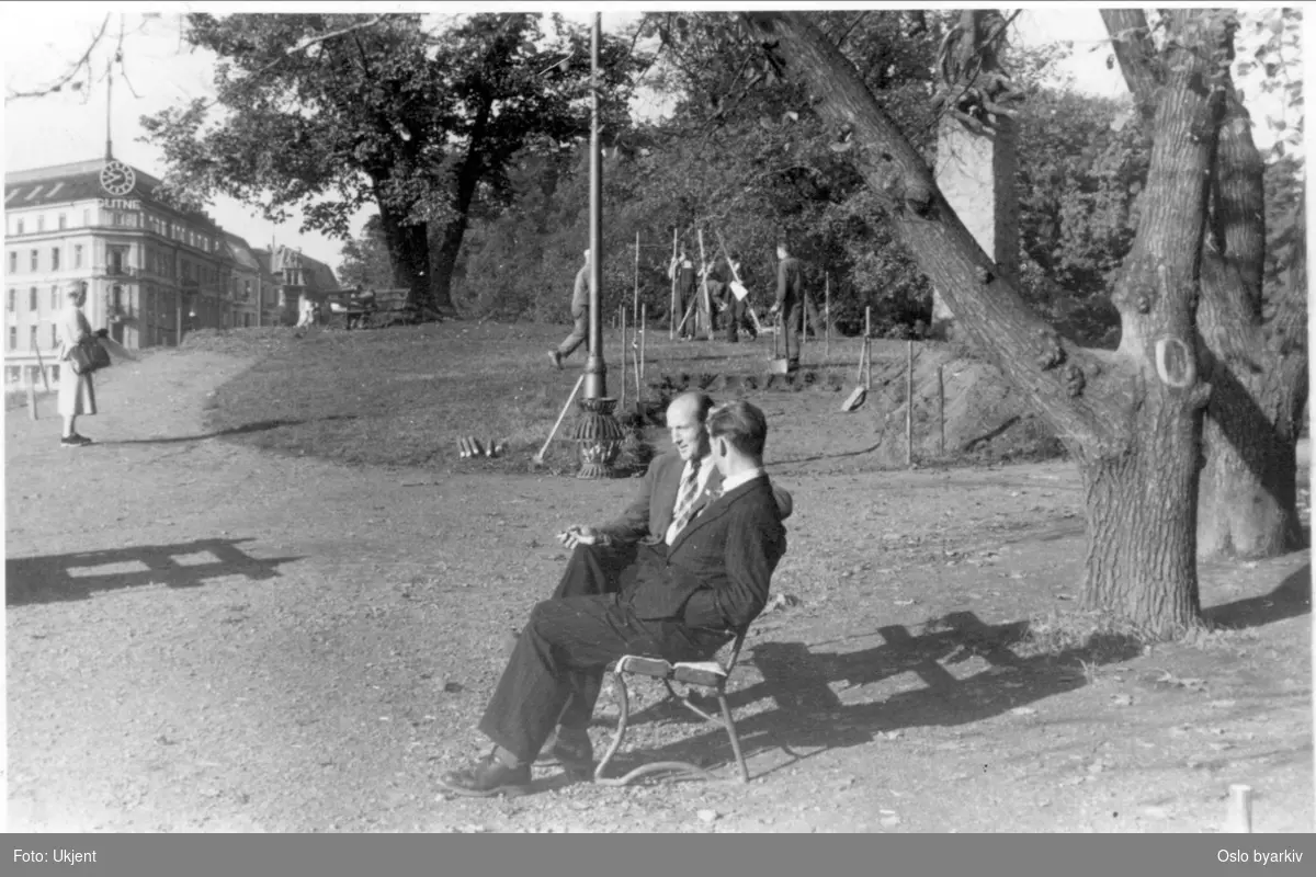 To herrer sittende på en benk ved Abelhaugen. Arbeidere i gang med oppmåling av gangvei over haugen ved Abelmonumentet. Kvinne iakttar arbeidet. Glitnegården i bakgrunnen. Bildet tatt 1949 - 50.