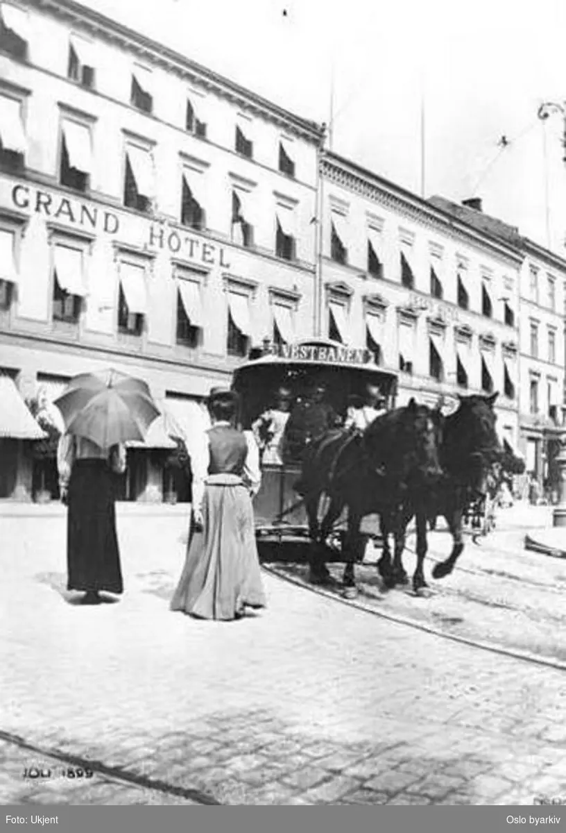 Tospanns hestesporvogn til Vestbanen svinger inn i Rosenkrantz' gate ved Grand Hotel, i juli 1899. Spaserende damer.