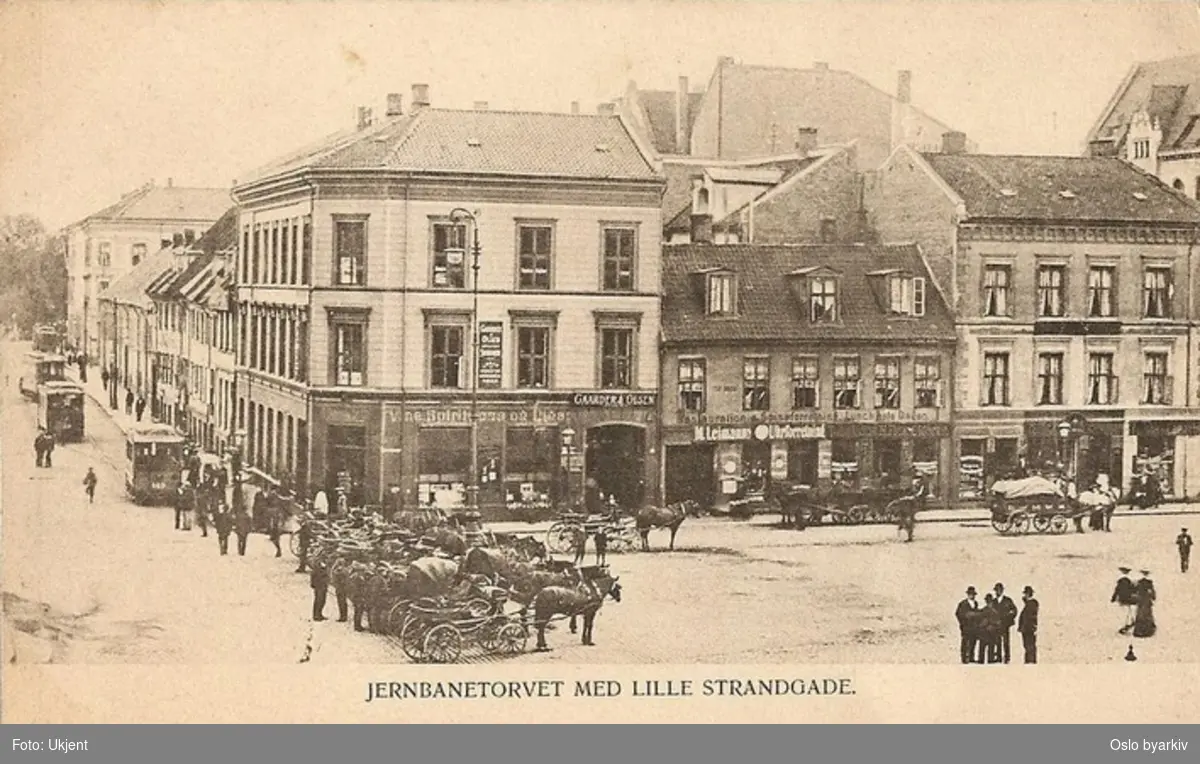 Jernbanetorget ved Lille Strandgate (Strandgata fra 1934) til venstre. Hestedrosjer oppstilt, hestetransport, butikker, Blåtrikker. Gamle bygårder med butikker der Amerikalinjens bygning står idag. Postkort.