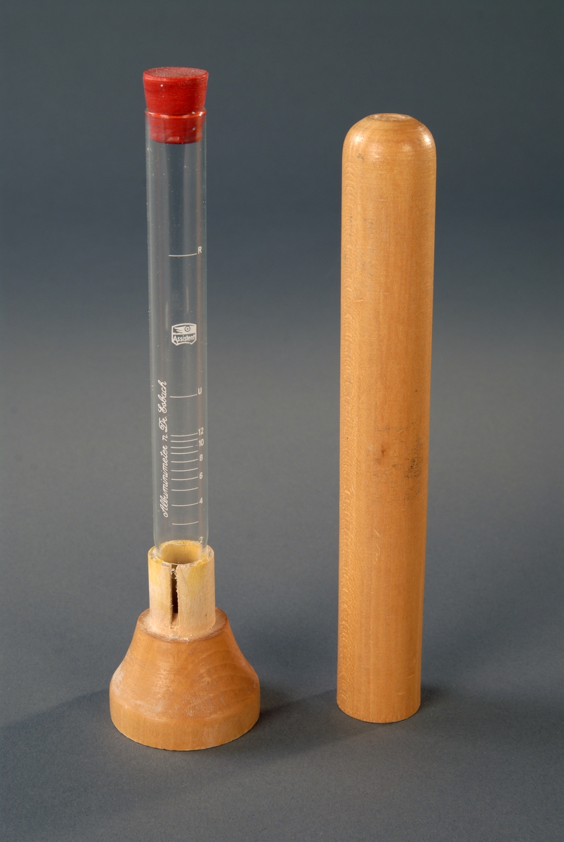 Et reagensglass m/gummikork stående i en tresokkel, med en sylindrisk trehylse over seg.