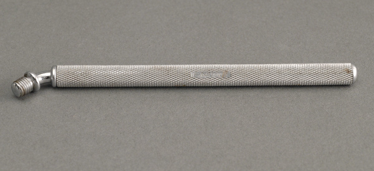 Sylindrisk metallbeholder, med ru overflate og en nålspiss stukket inn. Spissen kan i den ene enden trekkes ut, snues 180°, og monteres så den står i 45°.
