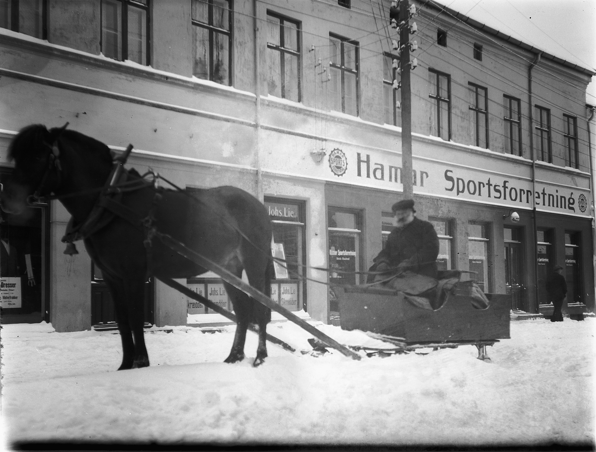 Hesten Trygg med Oluf Danielsen i slede i Torggata, foran Hamar Sportsforretning og Johs Lie Skrædder. Hamar. 