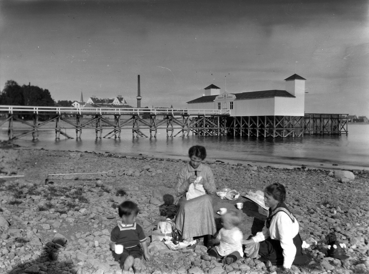 To kvinner og to barn på stranda ved Hamar badehus. Hamar Badehus ble bygget i 1916, og revet i 1931. Hamar Sjøbad.