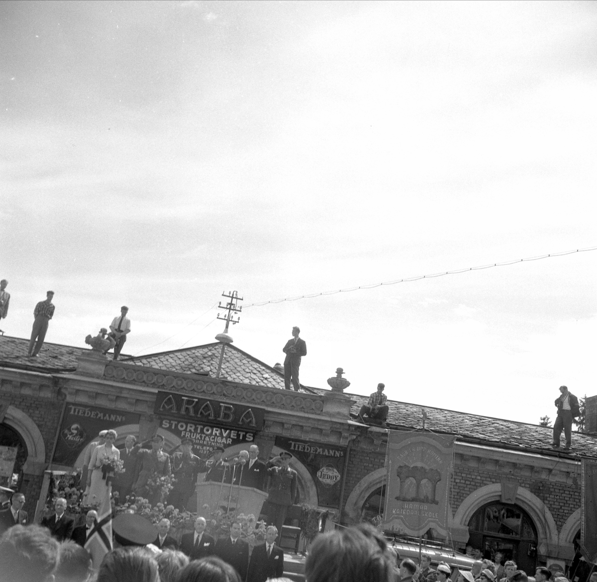 Hamar, Stortorget, signingsreisen 1958, kong Olav og prinsesse Astrid på en scene foran Basarbygningen, pressefotografer på taket,