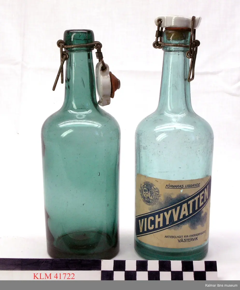 KLM 41722 Flaska, 6 st, glas. Flaska av genomsiktligt blågrönt glas, med patentkork av vitt porslin med gummiring och metallbeslag. Några av flaskorna har påklistrad etikett med blått tryck: Vichyvatten, Aktiebolaget nya centralbryggeriet, Västervik, Förvaras liggande.