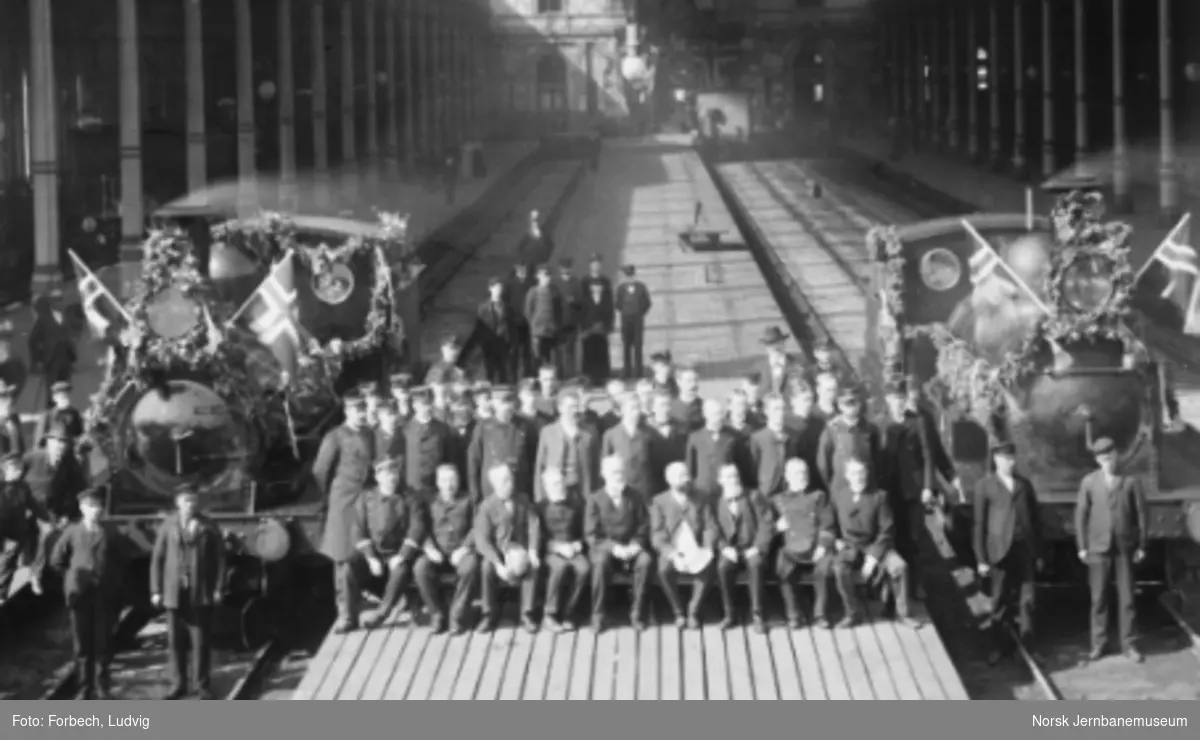 Stasjonspersonalet oppstilt på plattformen mellom to pyntede damplokomotiver i Østbanehallen ved Hovedbanens 50 års-jubileum