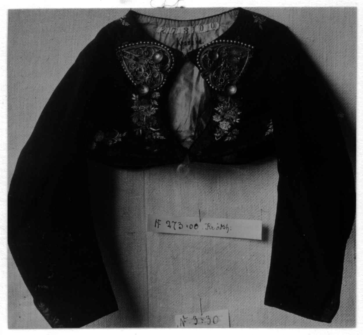 Svart trøye til jente av klede med dekor av svarte brosjerte silkebånd, blomster.
Trøyespenner av sølvfilligran (hører til NF.1900-0273 enl. referanser).