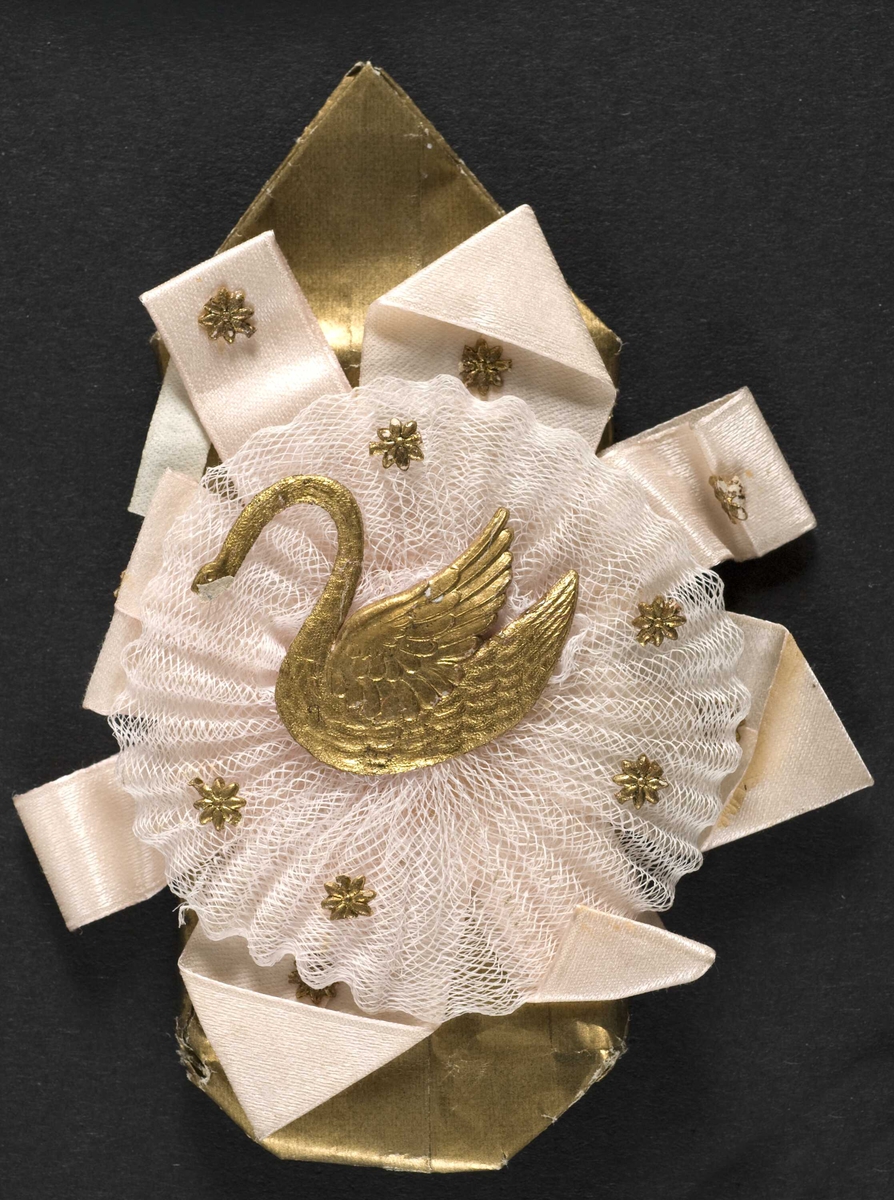 Pynt, dekorasjon laget av glanspapir, tyll og silkebånd. Forestiller en gullsvane på en rosett av lys rosa tyll.