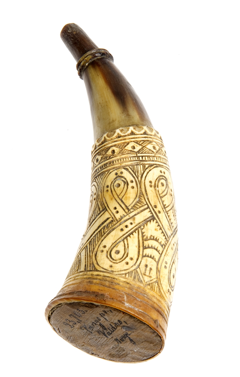 Krutthorn med flat bunnplate i tre. Ornamentikk på horn i form av horisontale bårder, noen tynne og en tykk som er fylt med et bånd som går i løkker kring hornet. Bakrunnen er også ornamentert med enklere ansikter som kikker frem mellom streker og buer.