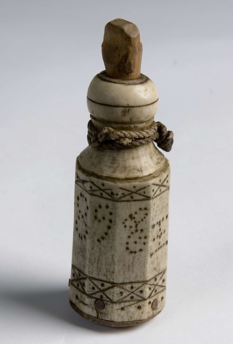 Ti-kantet flaske med kuleformet munning. Bunn og propp er laget av tre. Hyssing er festet ved halsen. Dekorert med kolrosingteknikk. 