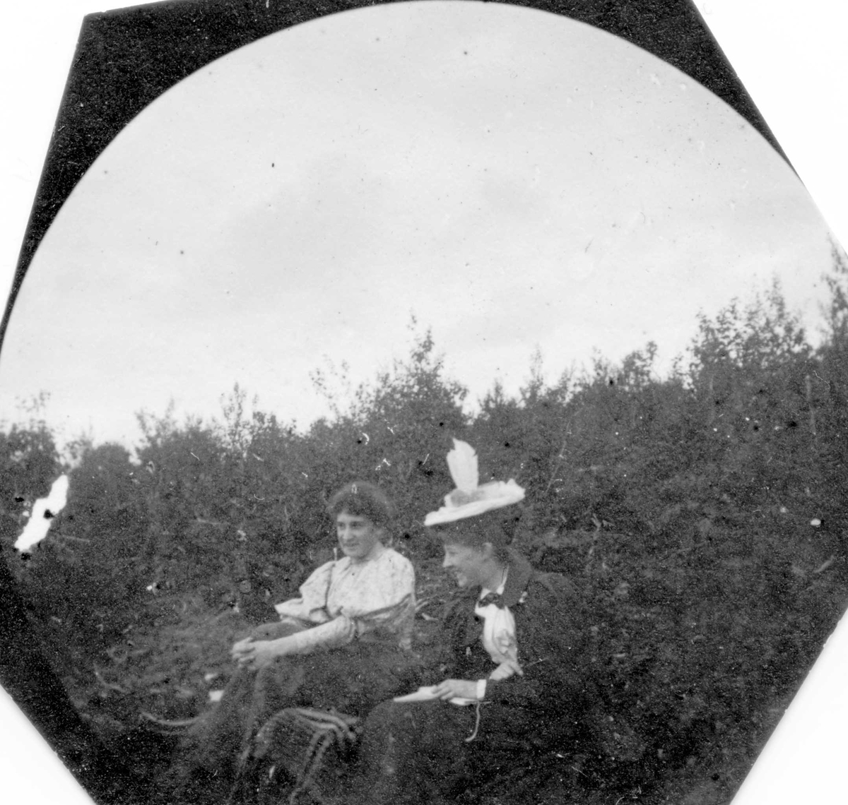 To damer sitter på bakken  i skogen, ser bort fra fotografen. En holder hendene rundt knærne.