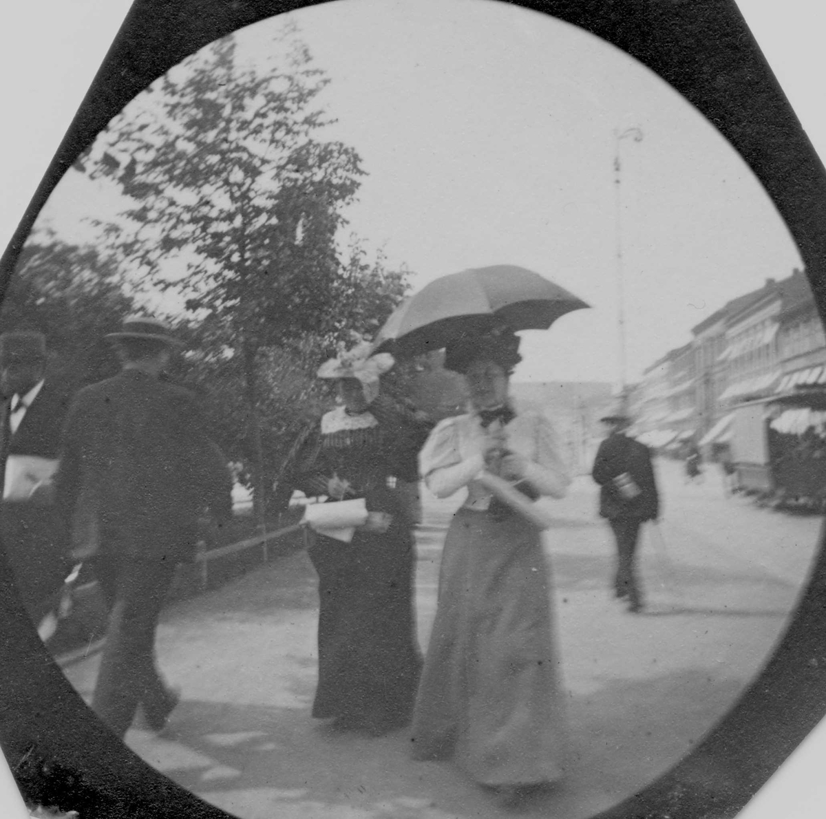 Mennesker spaserer på Karl Johans gate, Oslo. Kvinne med hvit jakke og paraply fremst i bildet.