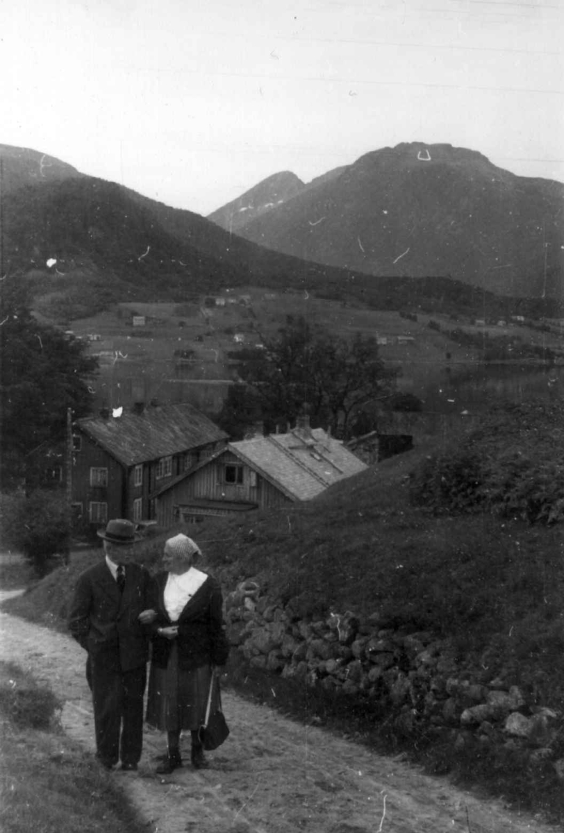 Daugstad gård i Tresfjord i Møre og Romsdal i 1954. Kvinne og mann går arm i arm på grusvei med bygda og fjellene i bakgrunnen, antakelig Skorgedalen.