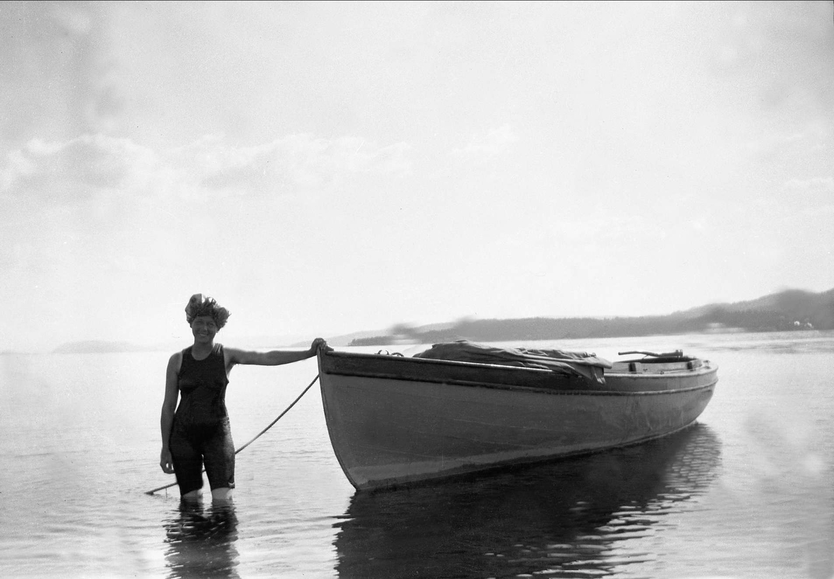 En kvinne i badedrakt poserer og holder i bauen på en robåt.Robsahm og Lund.