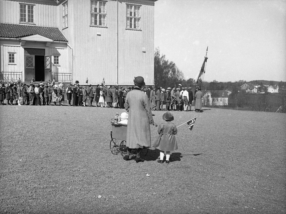 En mor med to barn står i skolegården 17. mai og ser på barnetoget går ut fra Bergan Skole, Nøtterøy. Fotografert 17. mai 1932.
