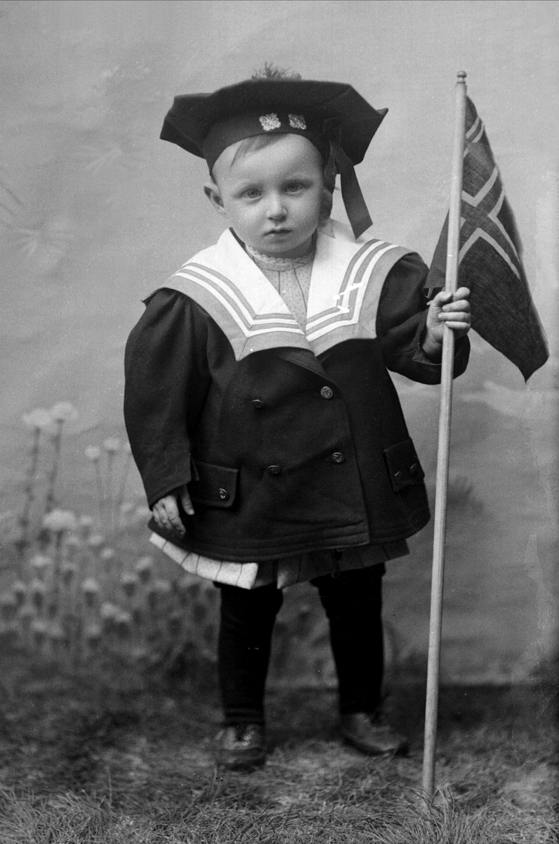 Portrett, liten gutt i matrosdrakt med det norsk flagget. Flagget er det norsk unionsflagget (1844-1899) men de svenske fargene er fjernet. Peter Gruner.