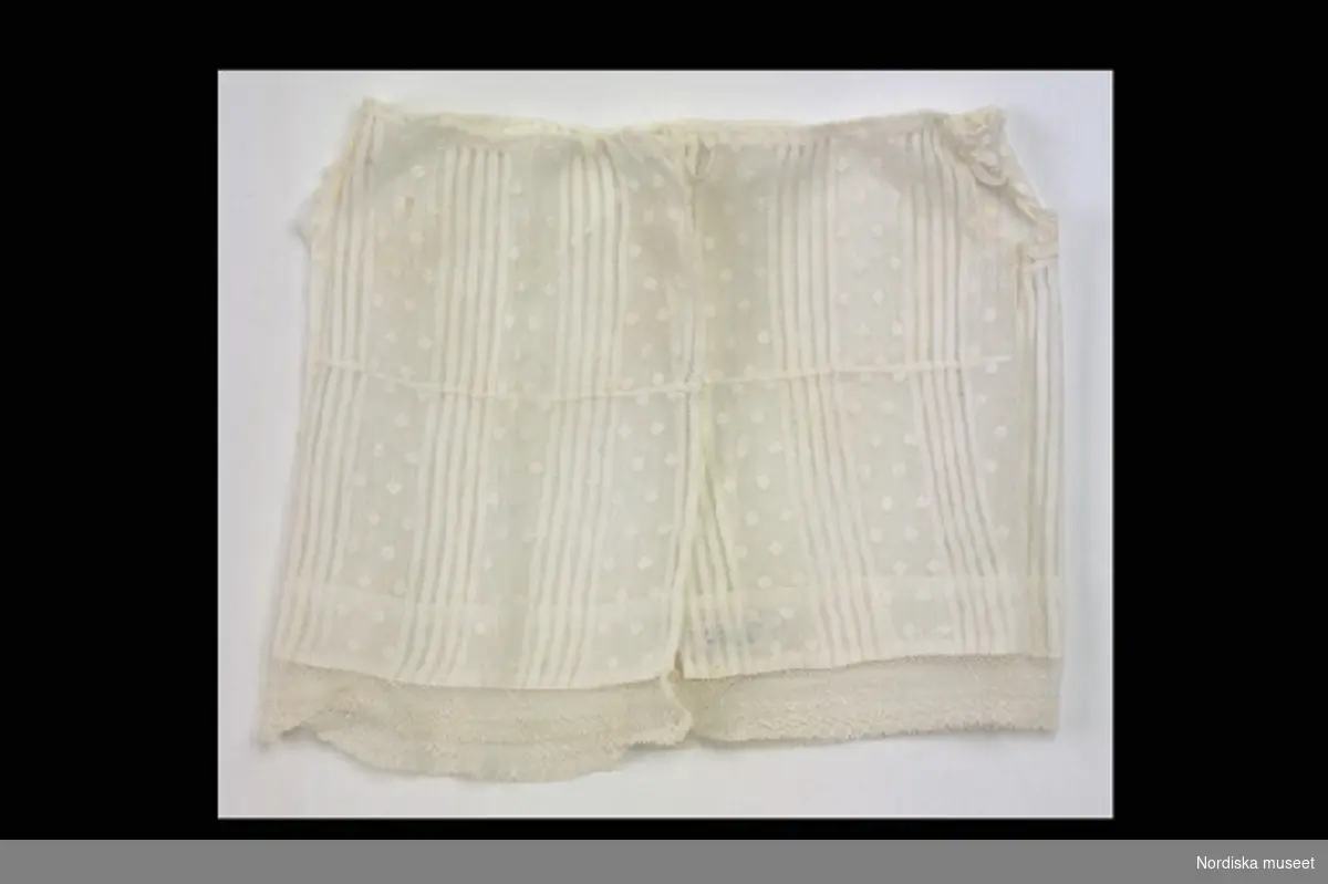 Inventering Sesam 1996-1999:
L 21 (cm)
Förkläde, dockförkläde, av vit bomullsvoile, omväxlande med genombrutna ränder och invävda prickar. Dragsko i halsen, spetskant nedtill  och kring ärmhålen.
Anna Womack 1996