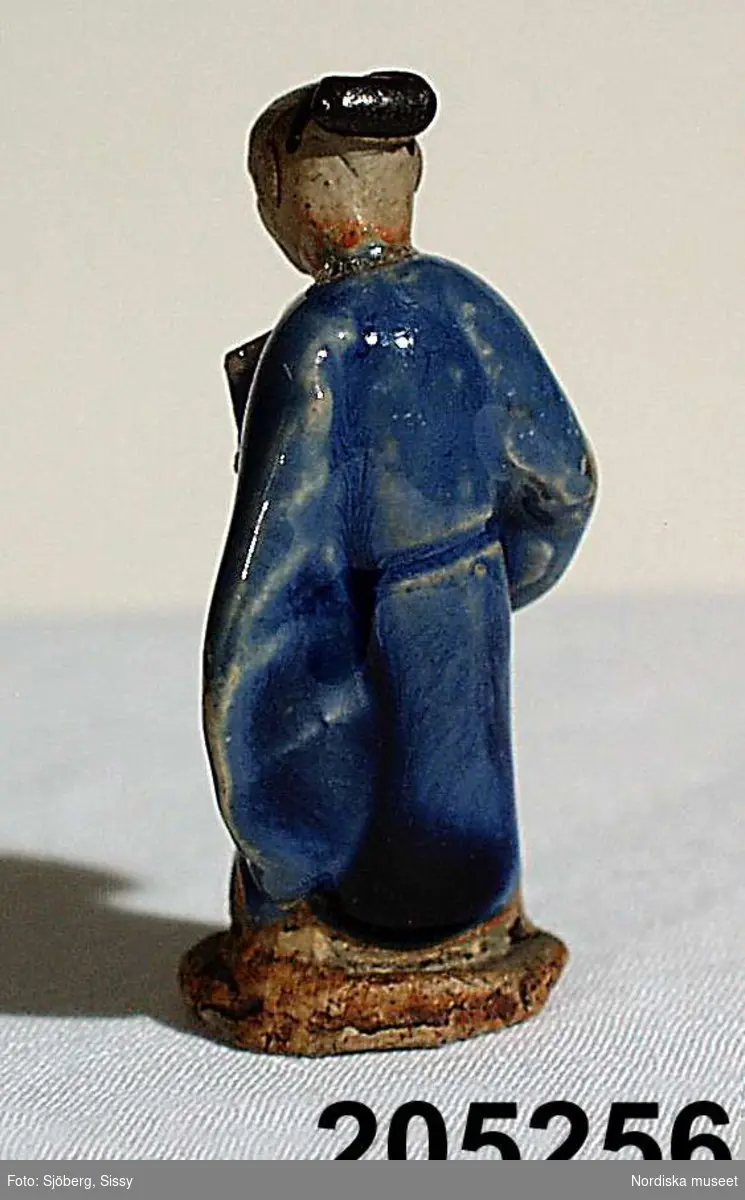 Stående figur klädd i blå dräkt, i ena handen en solfjäder. 