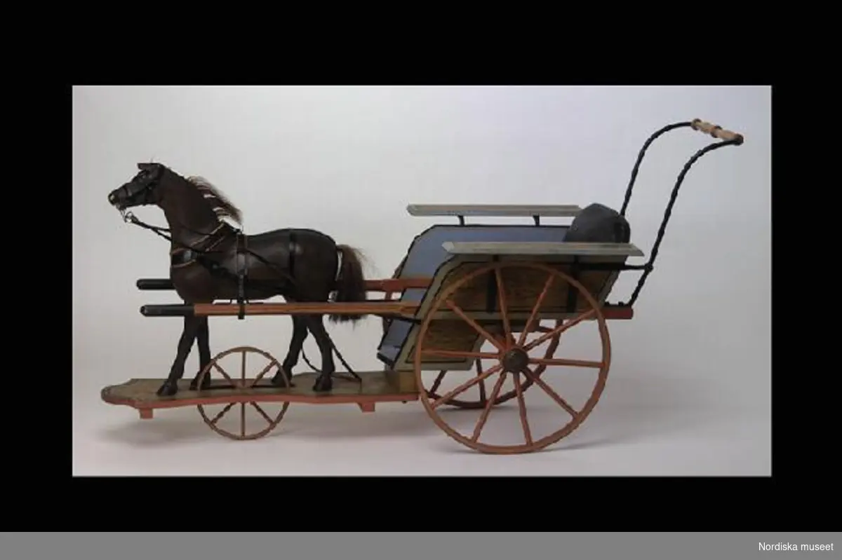 Inventering Sesam 1996-1999:
H  160   H  80  cm
Vagn förspänd av häst. Tvåhjulig vagn med läderklädd sits, ådringsmålade sidor och stänkskärmar, två hjul av trä skodda med järnband, baktill på vagnen utgår ett handtag. Häst av skulpterat trä, brunmålad , seldon, stående på ådringsmåkad fotplatta med ett hjul. 
Ekipaget kommer ifrån Villa Högudden, Lidingö, från f d sommarnöjet till dokor Ragnar Magnus Bruzelius.
Birgitta Martinius 1996