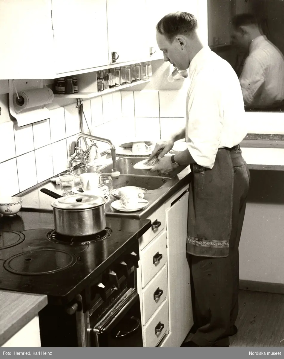 Kök. Interiör.  En man med förkläde står vid diskbänk och diskar. 