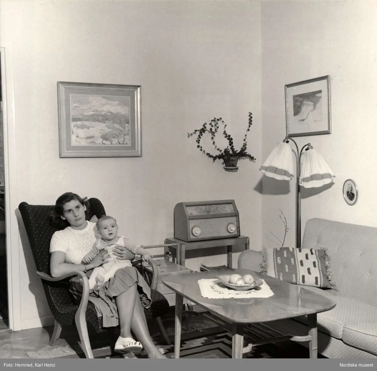 Vardagsrum, interiör. Kvinna sitter i fåtölj med litet barn i knäet. I bakgrunden en radio.