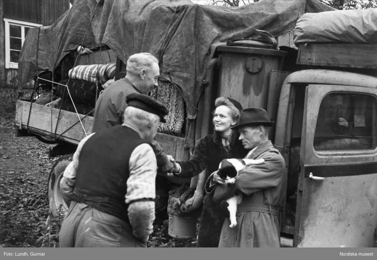 Statare. Paret Eriksson säger adjö till sina grannar vid en flytt. De står framför en lastbi, mannen håller en katt i famnenl.
