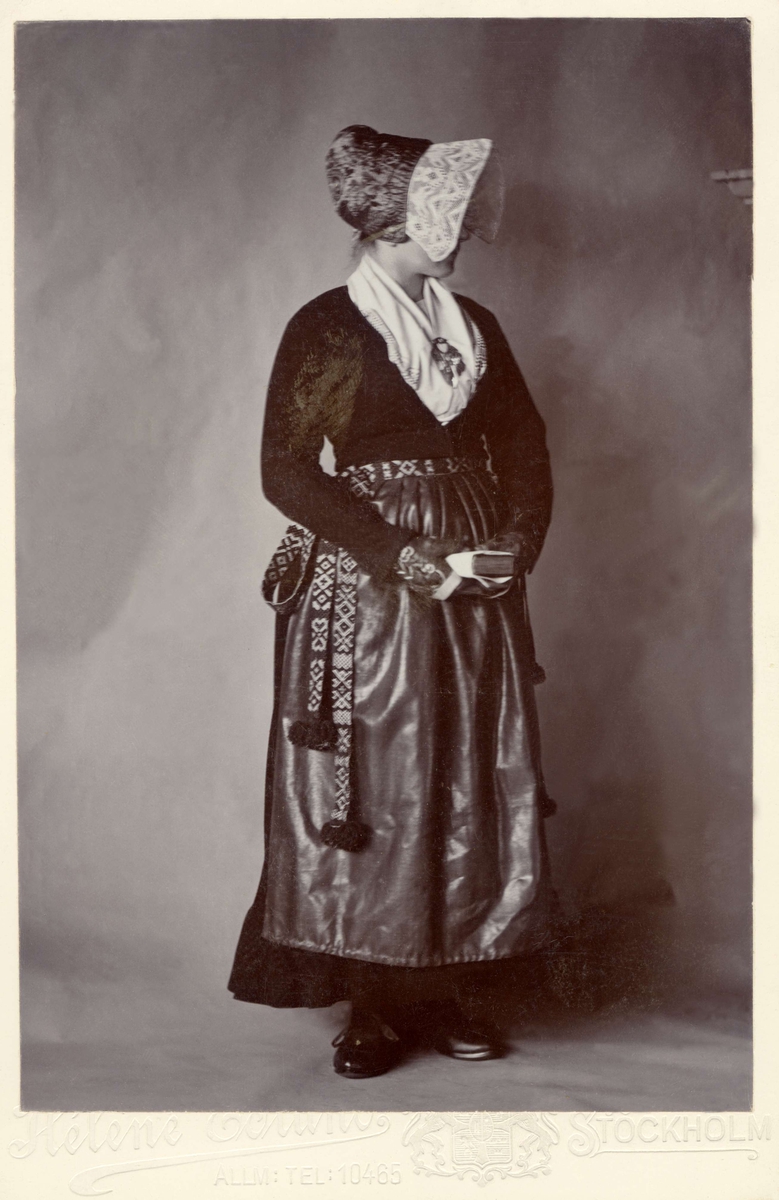 Porträtt av kvinna i folkdräkt från Dalarna, hon står med en bok i händerna, ansiktet bortvänt under hättan.