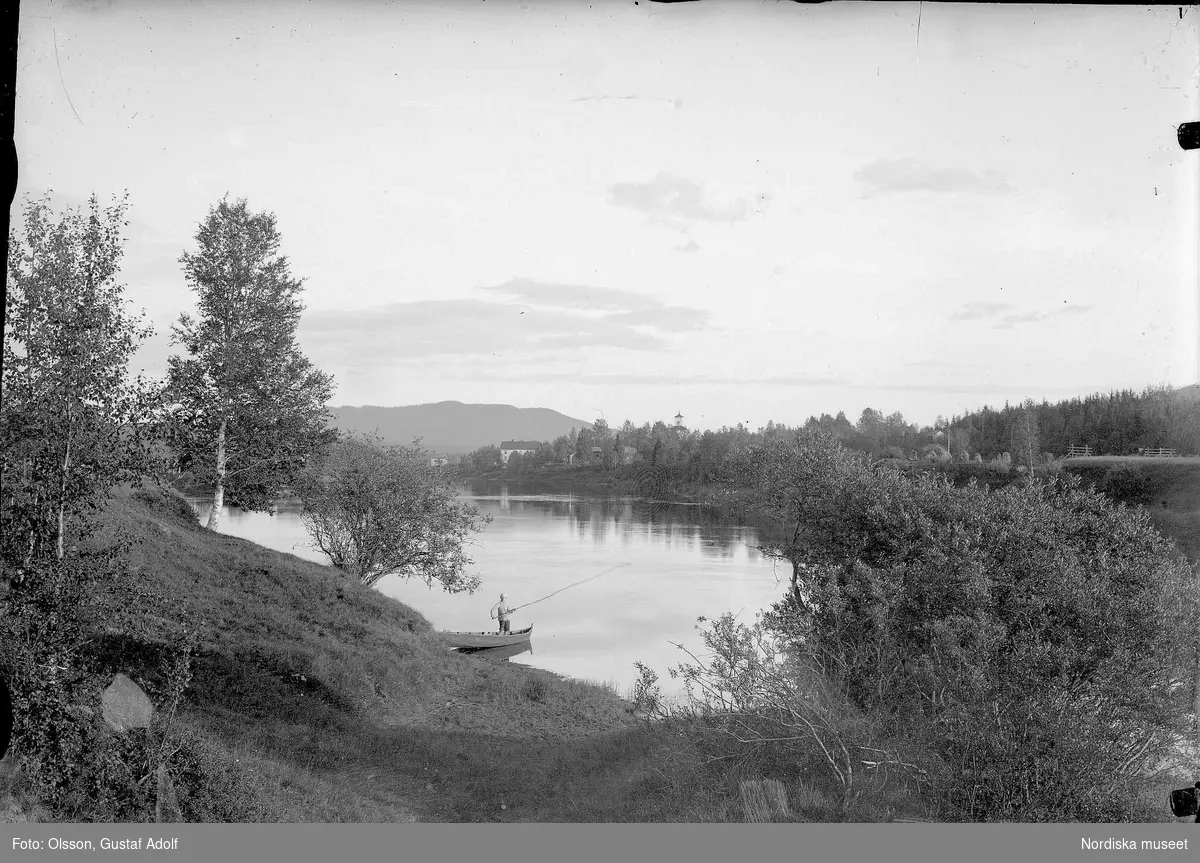 Utsikt över vattendrag. En man står i en båt  vid strandkanten och metar. Bild från början av 1900-talet.