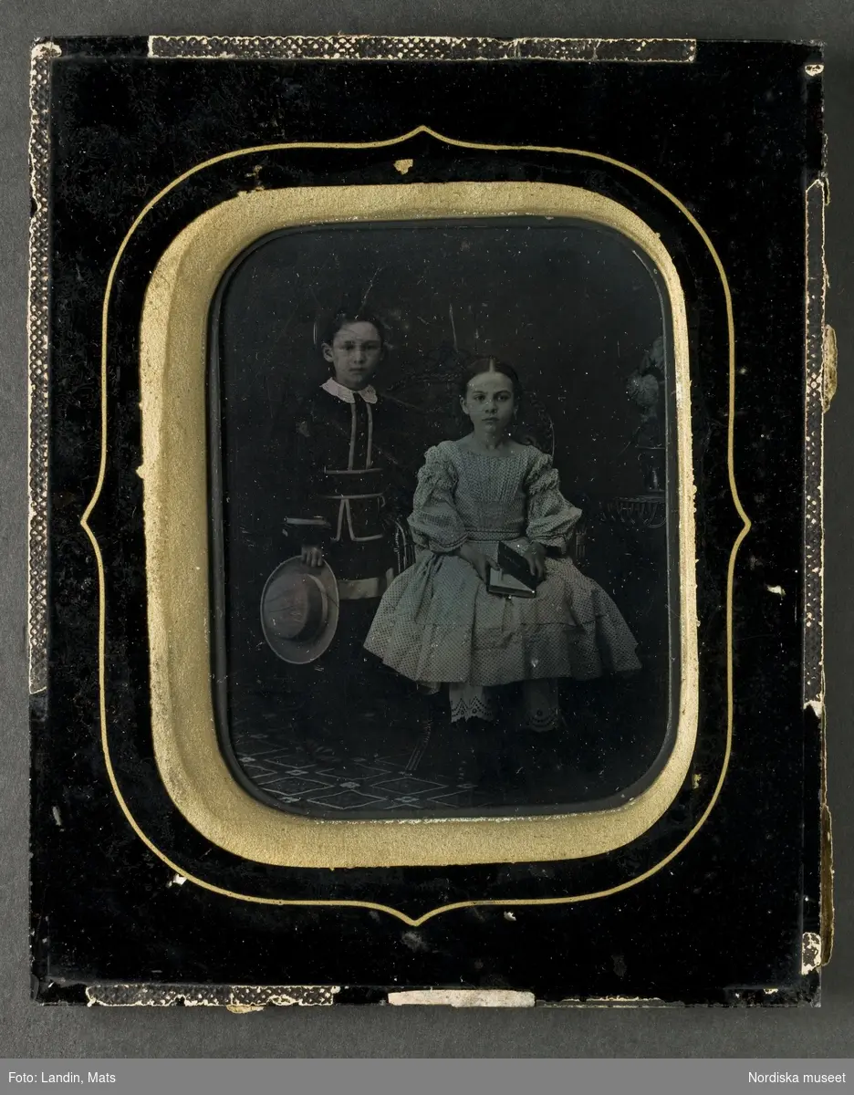 Barnporträtt, troligen två syskon, en pojke och en flicka i 6-7-årsåldern. Pojken, stående till vänster, klädd i svart/vit dräkt med vit krage. Håller en ljus hatt i höger hand. Flickan med mörkt mittbenat uppsatt hår, sittande i korgfåtölj, klädd i ljus klänning med mörka prickar och vita mamelucker. Håller en bok i handen. Ambrotyp, ca 1850-60. Nordiska museet inv.nr 231914.