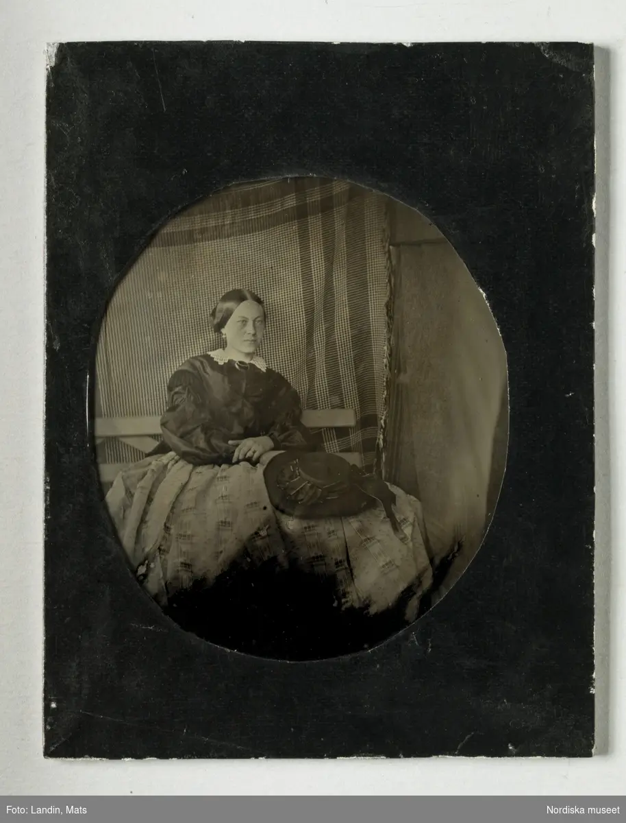 Damporträtt, helfigur. Sittande dam på bänk, rutig bordduk som bakgrund, i krinolin med stor hatt i knäet, ca 1860. Ambrotypi, fyrkantig glasplåt med ovalt snitt i ovansidans svarta papper. 
Nordiska museet inv.nr 314087.