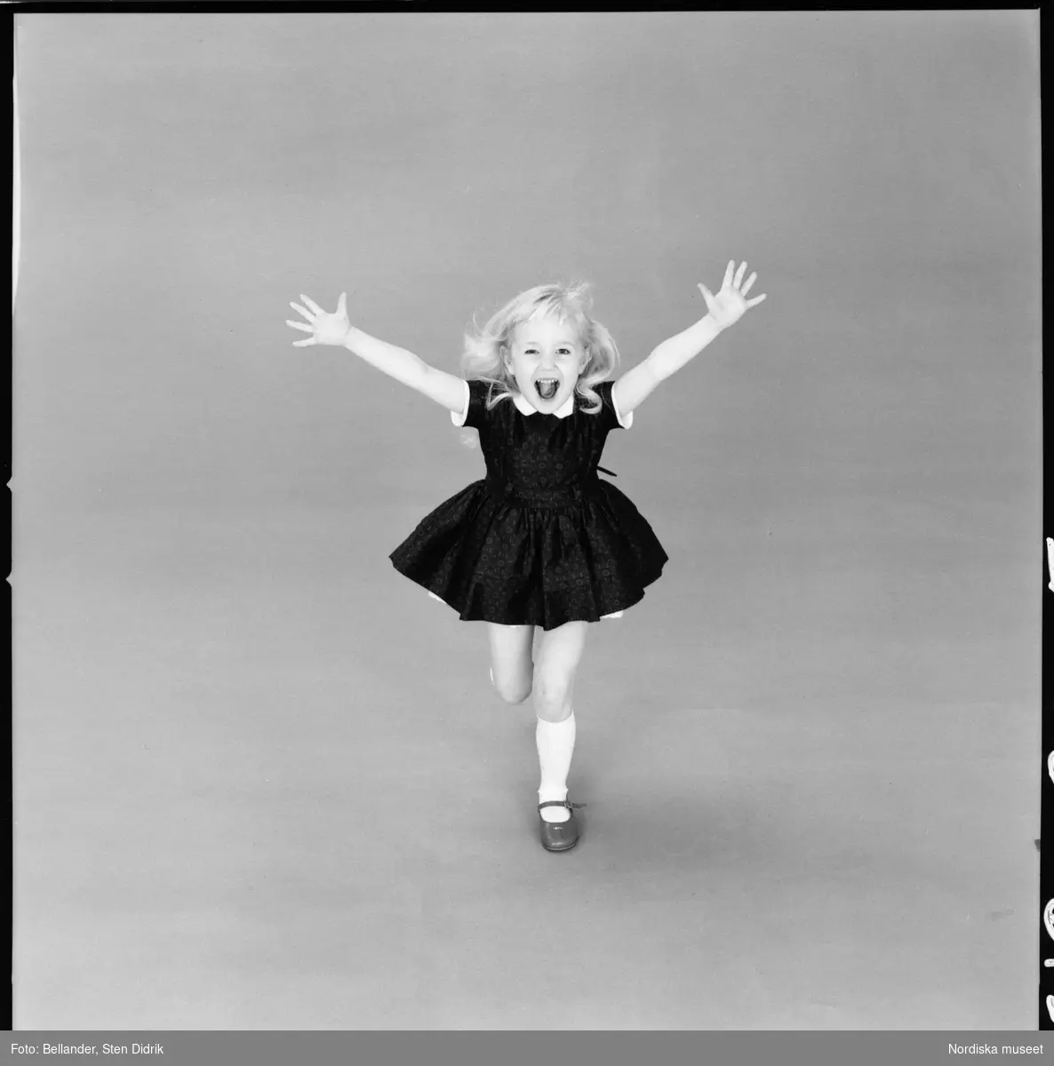 Barnporträtt. Flicka i klänning och skor med sleif springer med utsträckta armar mot kameran.