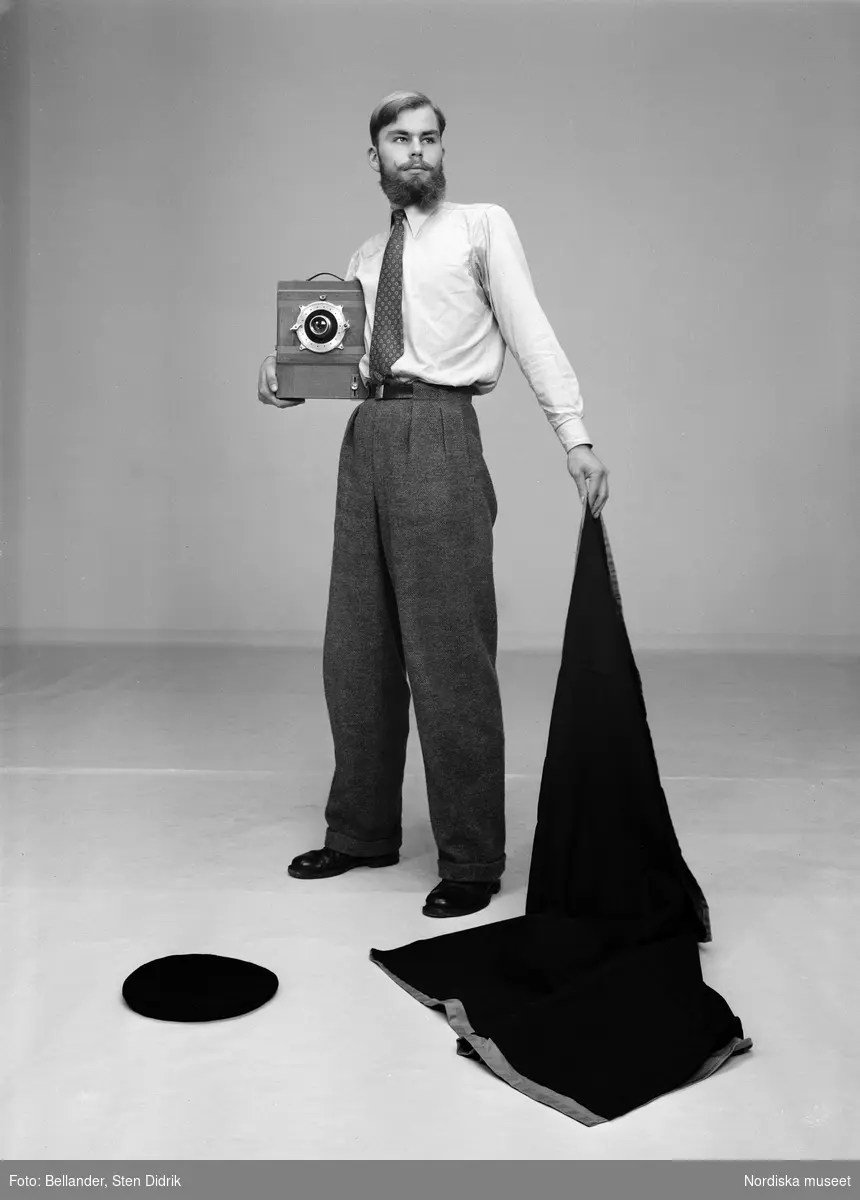 Fotograf Per Krafft håller kameraskynket som en slängkappa eller matadorskynke. På golvet ligger hans basker och i handen håller han storformatskameran.