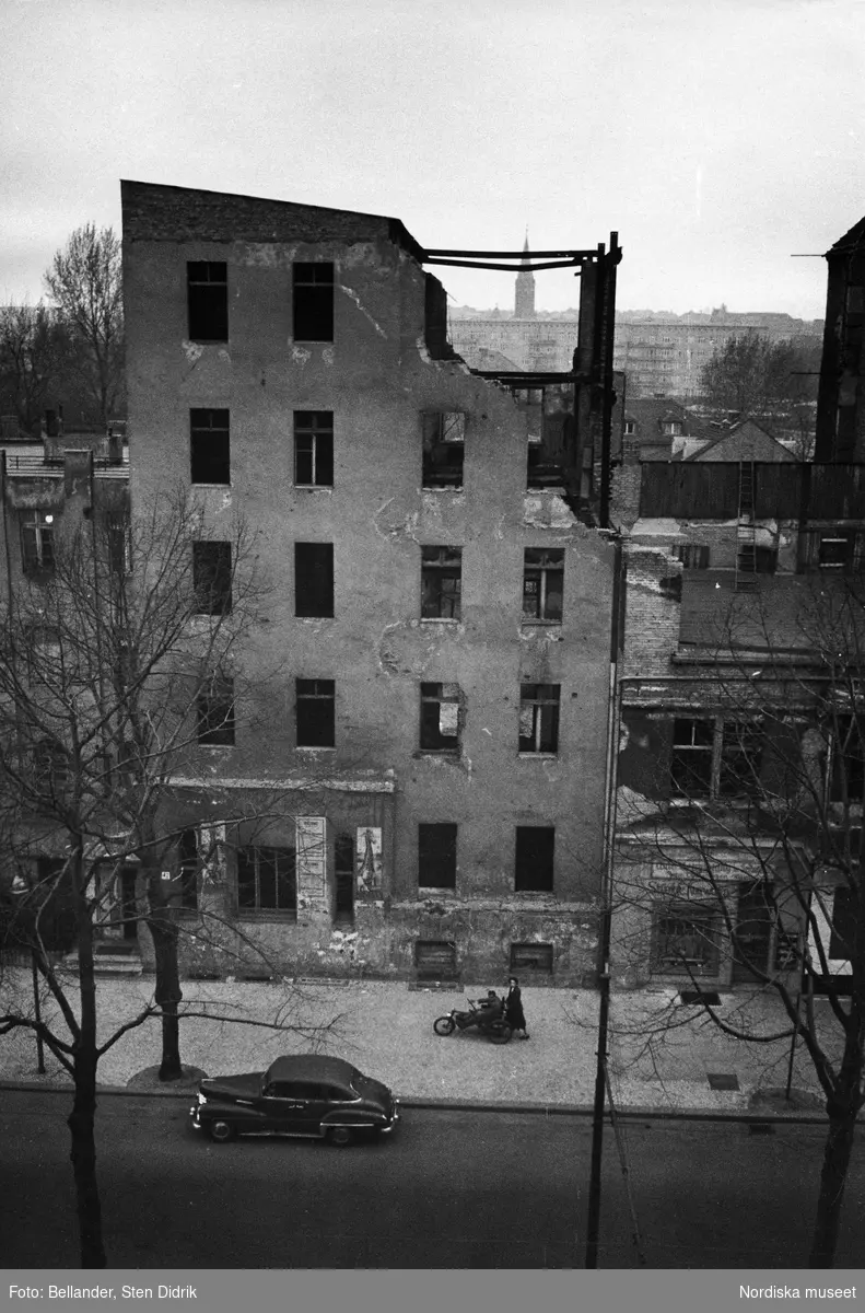 Utsikt från hotellrum. Vy över ett sönderbombat hus i efterkrigstidens Berlin. 
På gatan skjuter en kvinna en man i rullstol.