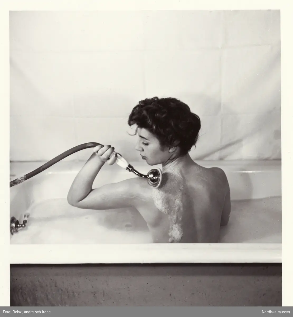 Badrumsinteriör. En mörkhårig kvinna sitter i ett badkar med badskum och duschar ryggen.