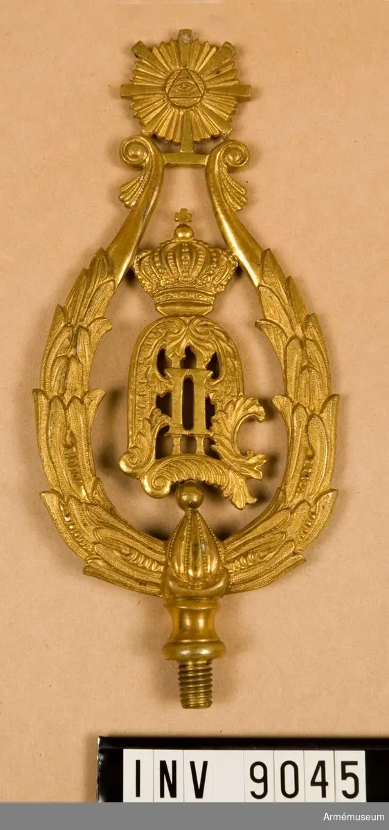 Spets O II av förgylld metall med konungens monogram omgivet av en krans, spetsformad. För fastsättning vid fanan en gängad skruv.