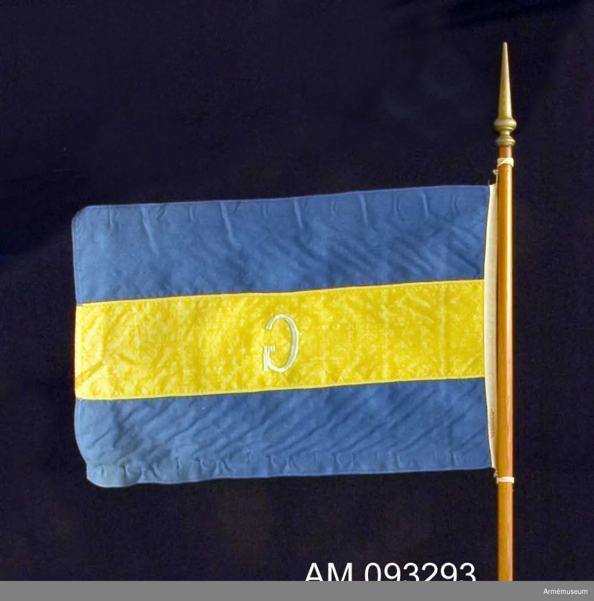 Enkel flagga i linne på trästång med svarvad knopp målad i guld. Utförd av Stockholms flaggfabrik AB.