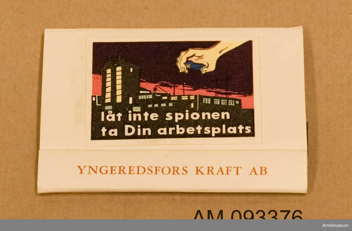 Tändsticksfodral med tändstickor. På ena sidan klistermärke med motiv som illustrerar hur en spionhand sträcker sig över en husbyggnad nattetid. 