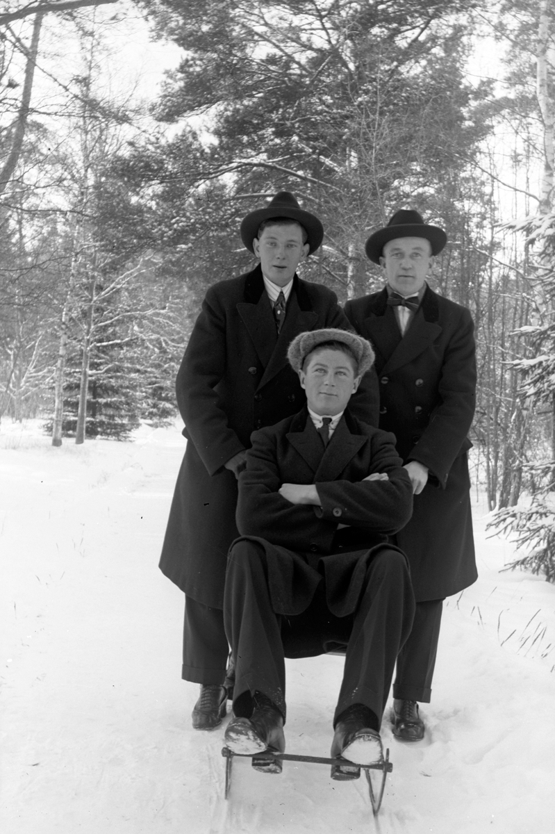 I en snöig skog sitter en man på en sparkstötting, två män står bakom honom.
