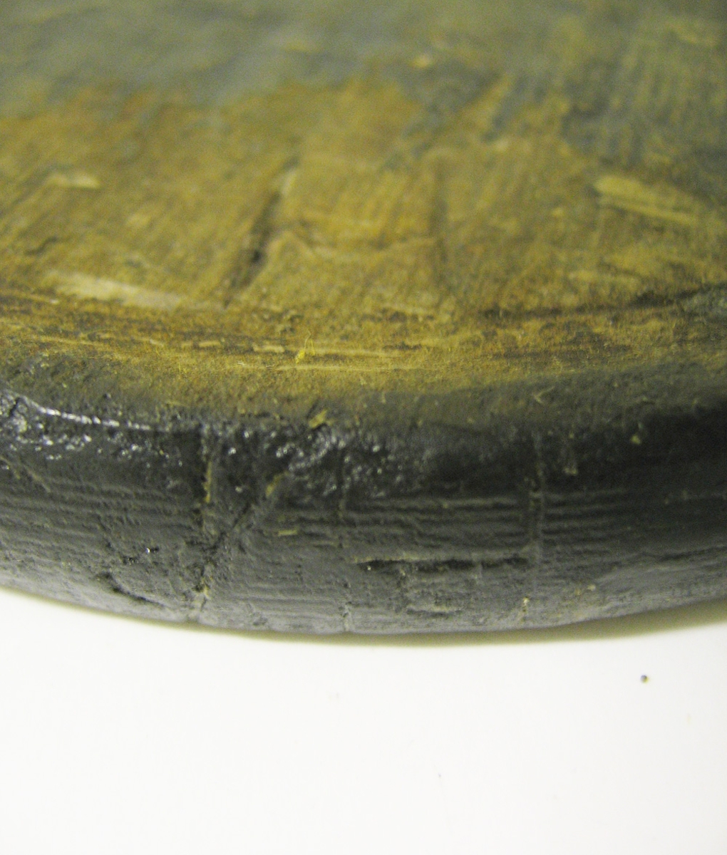 En svarvad disk av trä.
Disken är plan med upphöjda ytterkanter. På ena sidan av disken samt på den yttre kanten sitter två bomärken. På bägge sidor av disken finns tydliga märken i form av skärspår efter en kniv. På disken syns sex små hål.
Disken är välbevarad.

Text in English: A turned wooden platter.
A flat round disc with raised rim. On one side are two owner''s marks inscribed, one is located near the rim. Scored on both sides.
Well preserved except for six small holes.