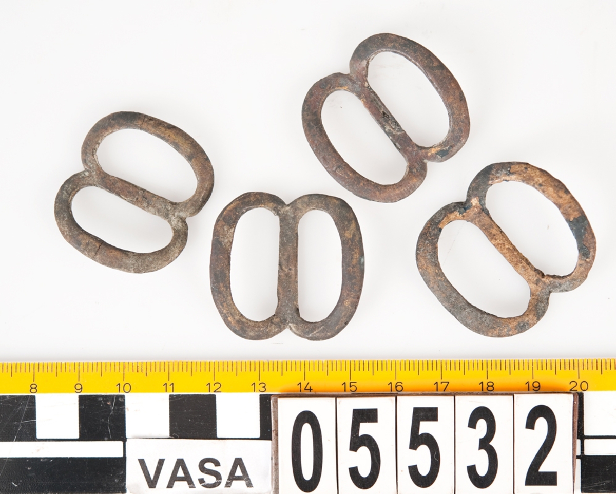 Fyra söljor av mässing. Mycket lika varandra med utformning som en liggande åtta. Ovansidan är kupig, undersidan är plan, något uppvikta från mittstolpen räknat.

Text in English: Four almost identical metal buckles.  One side of the metal strip is convex (probably the front), the other side is flat (probably the back).
The whole piece is concave, the center bar sits lower than the sides.  When viewed from the side, it appears to be a wide V.  From the front or back, it appears to be a flattened figure 8.
Each appears to be made by hand, seperately.  Potentially from one strip of metal, soldered in two places.