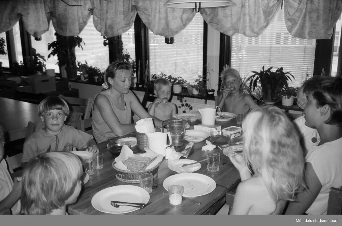 Vuxna och barn sitter vid köksbordet och har ätit mat. Porslin står kvar på bordet. Det verkar vara varmt ute för de har inte så mycket klädder på sig. Katrinebergs daghem, 1992.