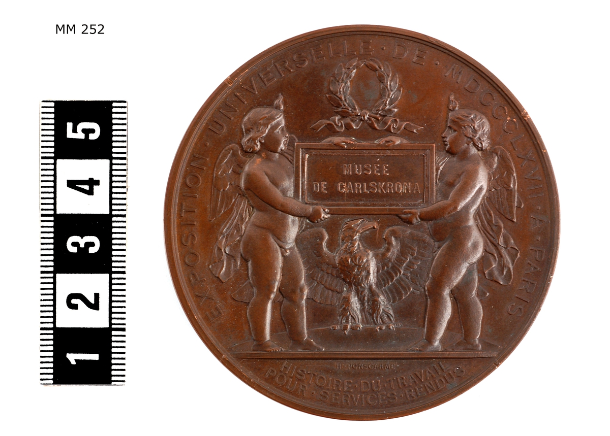 Medalj och diplom som tilldelats örlogsvarvets i Karlskrona utställning av fartygsmodeller mm vid utställningen i Paris år 1867. Medaljen i särskilt etui Diplomet inom ram av ek, polerad. Tillkom år 1868.