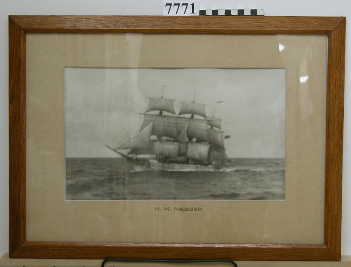 Tavla inom glas och ram av ek, fernissad.
Visar fotografi av skeppsgossebriggen Najaden under segel. Fotot inom grå kartong med texten: H.M. Najaden.
Neg.nr A 765 2:3