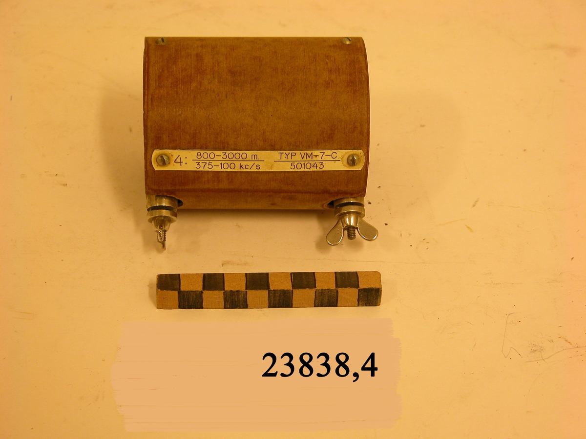 Ljusbrun cylinder med dubbla väggar och två skruvar av metall med vingmuttrar. På cylindern en fastskruvad bricka av plast med text : 4: 800-3000 m. 375-100 kc/s. TYP VM-7-C. 501043.