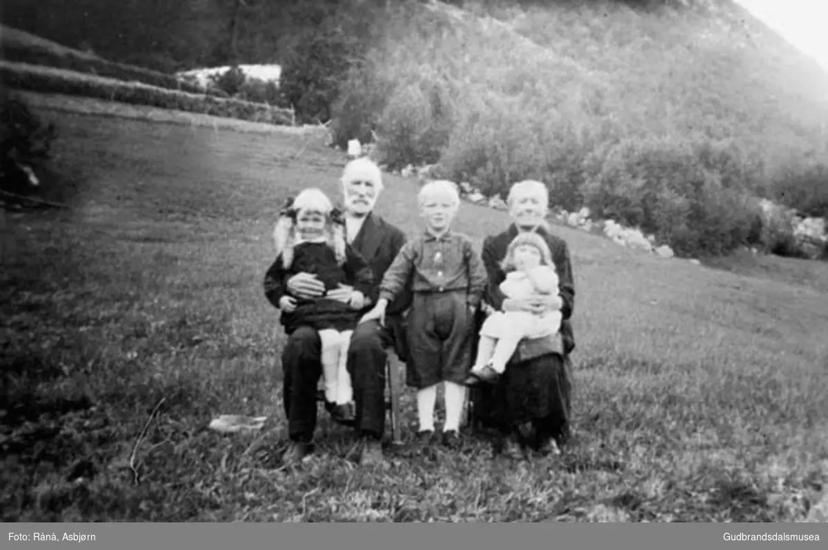 Familie med barn sitter ute, bildet er tatt på jordet i Nigard Rånå, fjell og hesjer i bakgrunnen.