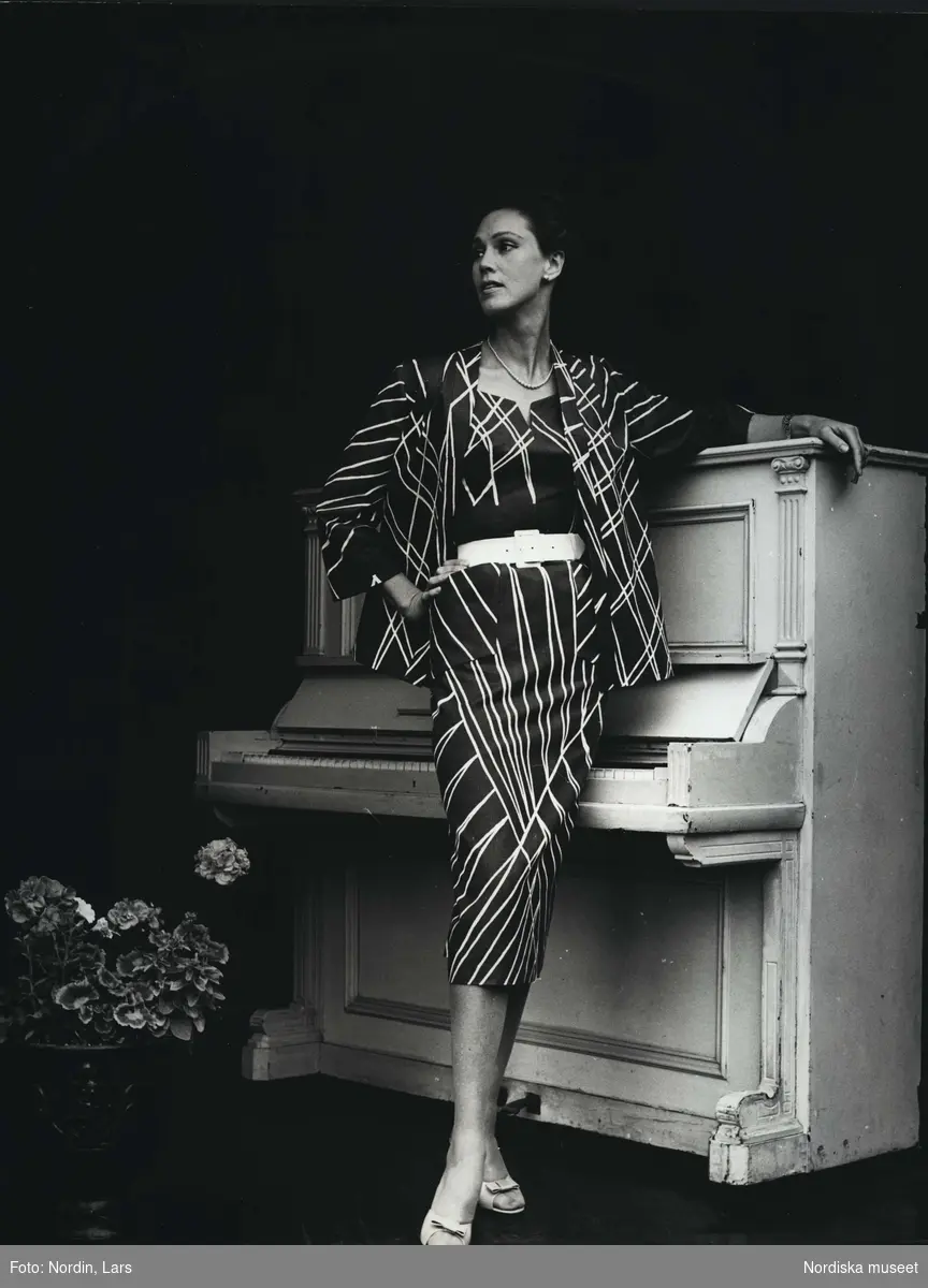Modell i klänning med jacka i mönstret "Scalino" designat av Viola Gråsten. Nordiska Kompaniets Textilkammare. Helsingborgsutställningen 1955. Modellen lutar sig mot ett piano. På golvet står en vas med blommor.
