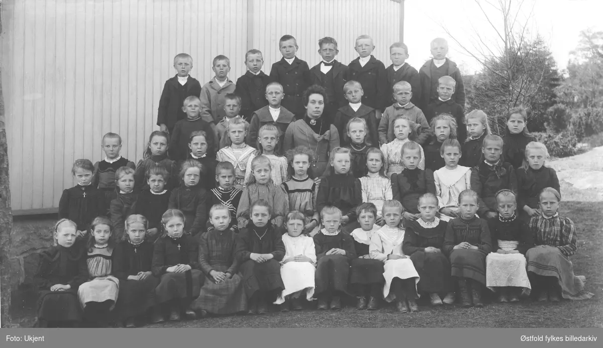 Nes skole, Rolvsøy ca. 1900. 
Første rad fra venstre: Signe Hauge, F. 1892. 
Skolen, i bakgrunnen,  blei seinere kommunelokale. Nes skole var forgjengeren til Rekustad skole nr. 2.