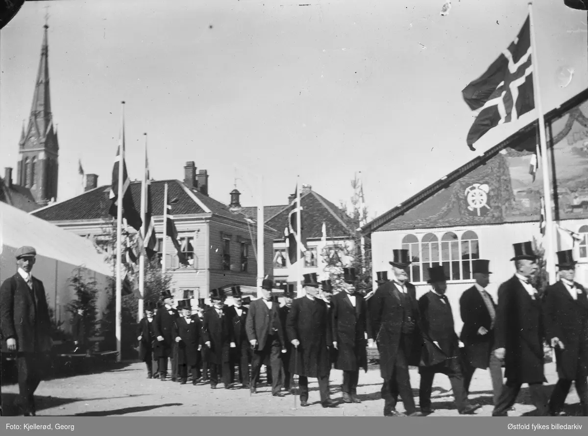 Engelsk besøk i  Arendal, ant. 1910.
Prosesjon/tog me menn i flosshat, falggborg. Trefoldighetskirken i bakgrunnen.